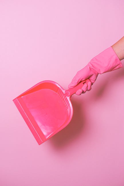 A hand wearing a pink rubber glove holding a pink dustpan against a light pink background, illustrating cleaning tools used in domestic or commercial surface cleaning and maintenance. The dustpan's su