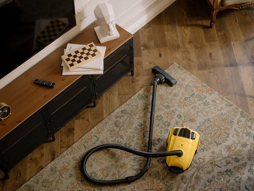 A person kneeling on a patterned carpet in a residential room, using a yellow portable vacuum cleaner for surface cleaning and deep cleaning purposes. The individual, wearing a beige jacket and blue j